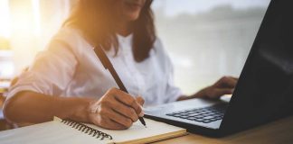 Person writing in notebook using laptop nearby