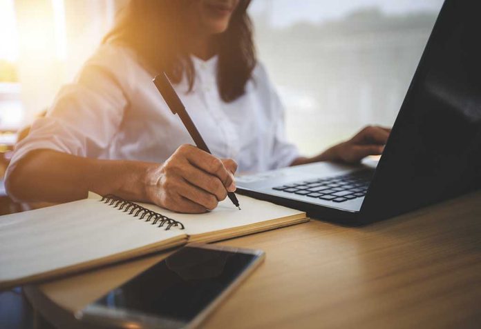 Person writing in notebook using laptop nearby