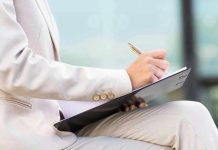 Person in beige suit writing on a clipboard.