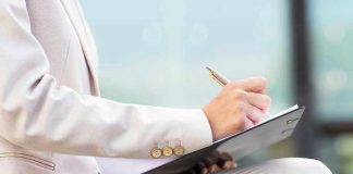Person in beige suit writing on a clipboard.