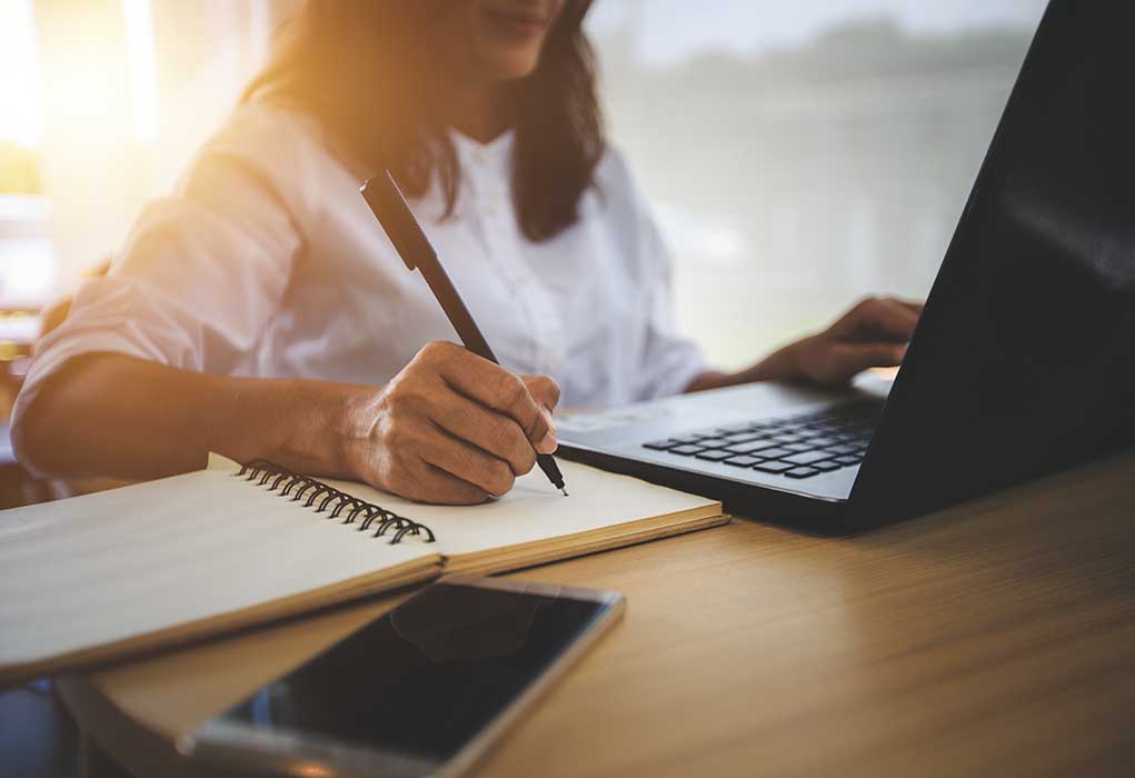 Person writing in notebook using laptop nearby