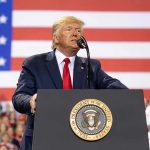 CAMPAIGN PLEDGE FULFILLED — Trump Signs Historic Order! Man speaks at podium with U.S. flag background.