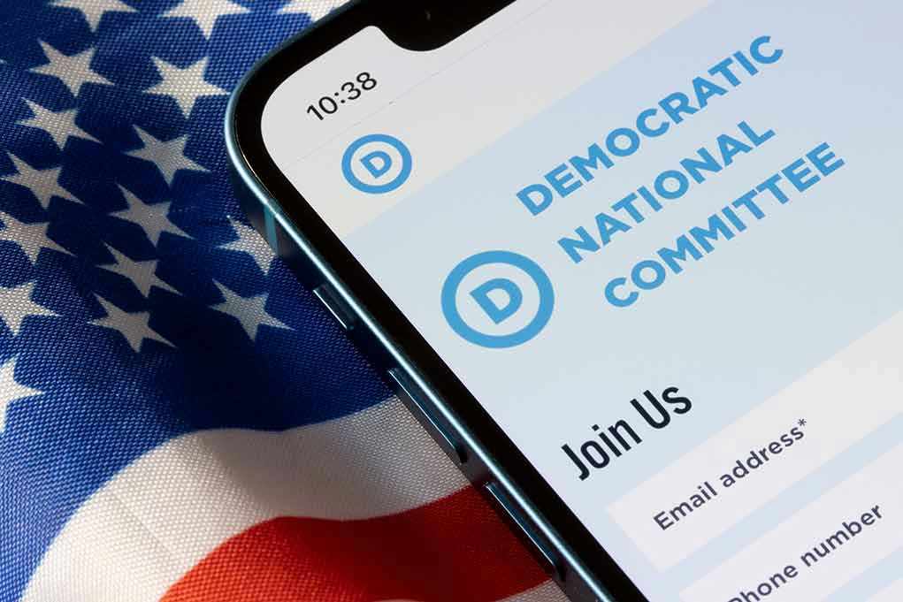 Democrats Panic as WORKING-CLASS REVOLT – They’re Scrambling! Phone displaying Democratic National Committee website over flag.