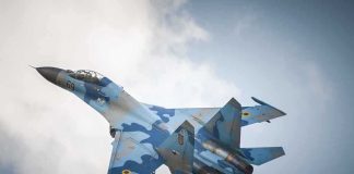 A blue camouflage fighter jet performing an aerial maneuver against a cloudy sky