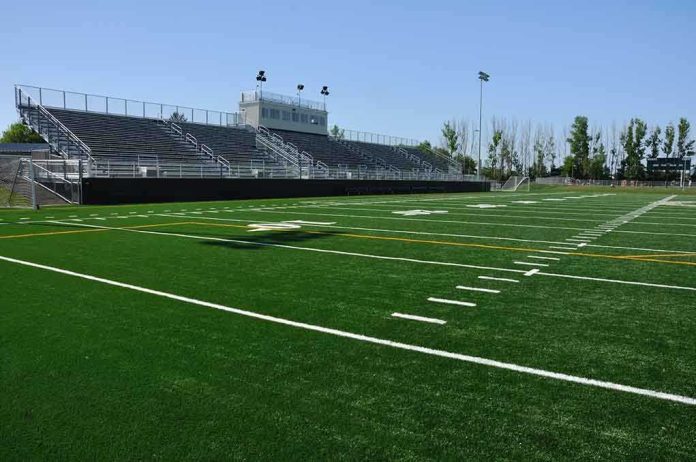 A football field with artificial turf and empty bleachers under a clear blue sky