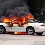 A white car engulfed in flames with thick black smoke rising