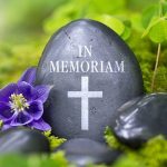 A memorial stone with In Memoriam and a cross, surrounded by flowers and greenery
