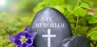 A memorial stone with In Memoriam and a cross, surrounded by flowers and greenery
