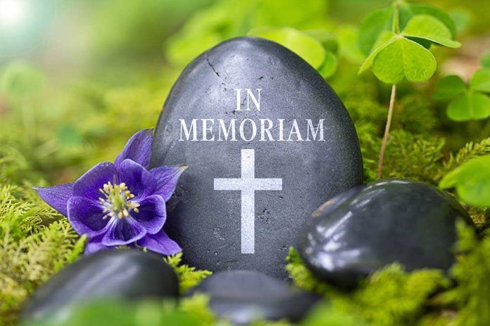A memorial stone with In Memoriam and a cross, surrounded by flowers and greenery
