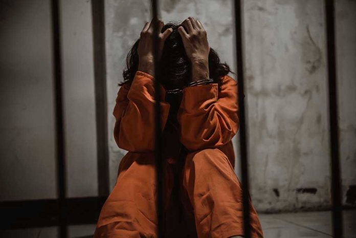 1484320151 Person in orange jumpsuit sitting behind bars, handcuffed.