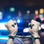 Hands in handcuffs in front of police cars.