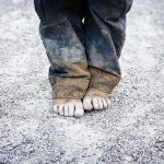 A child standing barefoot on a gravel surface with dirty pants