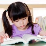 Young girl reading a book at a table with a bookshelf in the background