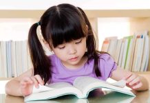 Young girl reading a book at a table with a bookshelf in the background