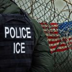 Police officer in tactical gear standing near a barbed wire fence with an American flag in the background