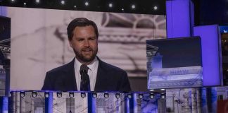 Man speaking at podium on stage, projection behind him.