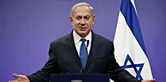 man in suit speaking at a podium with Israeli flag