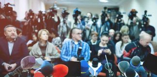 Row of microphones at a press conference with audience and cameras behind