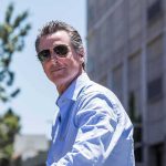 Migrants Gifted FREE College Ride – DOJ Steps In man in sunglasses wearing a light blue shirt outdoors