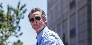Migrants Gifted FREE College Ride – DOJ Steps In man in sunglasses wearing a light blue shirt outdoors
