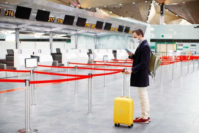 shutterstock_1684988143 Traveler wearing a mask stands by yellow suitcase at empty airport check-in