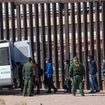 10,000 Seized – ICE Blitz STUNS! Border agents escort a group near a tall fence and a patrol van