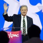 GOP Admits No Budget – Senator Sounds ALARM John Neely Kennedy waves while speaking at a podium on a brightly lit stage