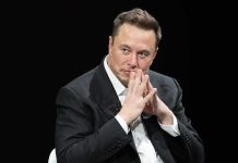 Elon Musk seated in a dark setting with hands steepled near his face