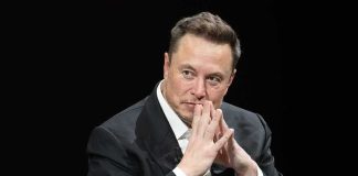 Elon Musk seated in a dark setting with hands steepled near his face
