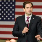 Marco Rubio speaking with a microphone in front of a large American flag
