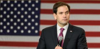 Marco Rubio speaking with a microphone in front of a large American flag