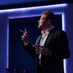 Grant Cardone EXPOSED In Class Action Lawsuit Man in suit giving a presentation on stage with blue backlighting
