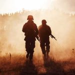 Two soldiers walking through a smoky field at sunset