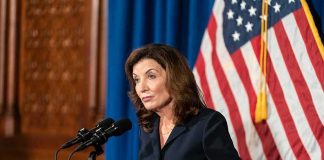 Hochul Weighs Ending $500M Medicaid Program After Fraud Raids Kathy Hochul speaking at a podium with microphones and a U.S. flag behind her